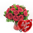 A dramatic Valentine's-style arrangement is presented against a pristine white background, featuring a lush bouquet of red roses in a ruby glass vase paired with a gleaming heart balloon. The bouquet consists of roughly a dozen or more deep crimson roses, their petals thick, velvety, and sensuously curved. Some flowers are fully open, showcasing layered whorls that create a sense of fullness and luxury, while others remain partly closed, their tighter centres hinting at feelings still blooming. Nestled among the roses is a generous amount of glossy green foliage, whose smooth, deep emerald leaves provide a cool, refreshing counterpoint to the fiery red tones. The vase is crafted from translucent red glass with subtle vertical fluting; the ridges catch the light and create slender highlights that run down its sides, while the stems inside are muted by the richly coloured water. In the foreground to the right, overlapping the vase slightly, floats a brilliant metallic red heart-shaped foil balloon. The balloon has a mirror-like finish that reflects the brightness of the setting, and stretched across its centre is the message I Love You, written in bold, flowing white script that stands out clearly. A stylised silver arrow, recalling Cupid's traditional symbol, appears to pierce the heart diagonally from lower left to upper right, reinforcing the romantic sentiment. Together, the roses, vase, and balloon form a classic declaration of affection, the sort of striking gift that might be ordered for same-day delivery around Brent Cross, Neasden, or along the North Circular to celebrate anniversaries, Valentine's Day, proposals, or any heartfelt moment when words alone are not enough.