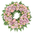 In this image, a lush, circular sympathy wreath is seen from above, positioned on a clean, bright white surface that lets the colours and textures of the flowers speak clearly. The wreath's shape is defined by a thick underlayer of dark green fern fronds, their delicate, feathery structure forming a soft, slightly ruffled border around the entire ring. Rising from this foliage, large roses in muted shades of dusty lavender, antique mauve, and softened greyish-purple are positioned with deliberate care. Each bloom is fully open, revealing a generous mass of tightly layered petals that curl gently outward, giving the arrangement a feeling of fullness and calm. Threaded among these larger roses are smaller blush-pink spray roses and dainty rosebuds, some just beginning to open. Their tender pink hue lightens the cooler purples and introduces a subtle warmth that feels comforting, like a quiet word of support. Tiny infills of darker greenery and minute buds sit tucked in the spaces between the flowers, deepening the shadows and adding subtle visual interest without breaking the wreath's serene rhythm. The neutral white background and soft, even lighting evoke the impression of a peaceful florist's workspace near Brent Cross station, where the arrangement has just been completed. The overall mood is one of respectful elegance and gentle solace, making this wreath particularly suitable for a funeral, memorial, or celebration of life service in and around Brent Cross, offering a graceful floral expression of sympathy, remembrance, and enduring love.
