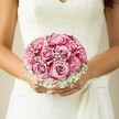 There's a poetic stillness in this image-a bride from Brent Cross ushering in her special day, her bouquet glowing against the canvas of an elegant white gown. Soft-petaled roses, numbering near a dozen, bloom from gentle, buttery pink at the center to bright, confident magenta at their fringes-each a testament to thoughtful arrangement. Threads of white baby's breath spiral throughout, their airy clusters imparting a sense of innocence and gentle anticipation, like a veil of countryside mist winding through the blossoms. The bride's gracefully tanned arms, finished with dainty, pale-shimmer nails, cradle the flowers with a warmth that hints at loving memories made on local avenues. Over her sleeveless lace bodice, a delicate sash gathers softly, the fabric's movement suggesting preparation and excitement. The surrounding backdrop, softly faded to warm beige, ensures all attention lingers on the vibrancy and tenderness of the crafted bouquet. From hand to petal, the arrangement expresses the artistry and emotion cherished in Brent Cross ceremonies, celebrating the transformative beauty of wedding flowers and the cherished bonds they honour.