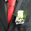 Set against the smart charcoal of a tailored jacket, an ivory rose boutonnière takes centre stage, its velvety petals catching soft daylight and suggesting a steady, refined warmth beneath a cool exterior. Tiny sprays of baby's breath bracket the bloom like a misty veil, their pristine dots bringing a note of innocence and lift, while slender slivers of fern and twisted leaves arc away with graceful motion to introduce depth and a dark-green contrast. The ensemble is punctuated by the tactile sheen of a red silk tie and the subtle texture of a checked shirt visible nearby, elements that hint at a groom or a guest dressing for a momentous occasion. A narrow white ribbon forms a small, neat bow at the base, completing the composition with an understated ceremonial flourish. The bouquet is fresh to the touch, faintly perfumed with the quiet, honeyed scent of rose and the green, almost herbal note of foliage. As Brent Cross Flowers, we craft pieces like this for weddings and special events around Brent Cross Shopping Centre and Cricklewood Broadway, hand-assembling each boutonnière with care so it remains secure and pristine from the florist's bench to the vows or reception. The result is classic, intimate elegance-a simple floral statement that reads as commitment and calm.