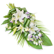 A long, graceful funeral spray of white lilies and lisianthus stretches diagonally across a white background, its shape flowing smoothly from a rounded, lush top to a fine, tapering end. The principal flowers are oriental lilies in immaculate white; some blossoms are already open wide, with their broad petals gently arching back to reveal pale green throats and light stamens, while several buds remain closed, their elongated forms adding height and quiet expectancy. Threaded among these are clusters of lisianthus, whose smaller, bell-shaped flowers bring a soft, ruffled texture. Many of the lisianthus petals are edged with a subtle lavender hue, creating a delicate halo of colour that sits peacefully against the prevailing white and green. Numerous buds dot the design, catching the eye and adding dimension. The foliage underpinning the spray is varied and carefully placed: broad, dark green leaves support the blooms like a calm, protective layer, and some leaves have been artfully rolled or folded into neat, cylindrical forms that contribute an architectural interest. From one side and along the lower portion, thin, palm-like fronds extend outward, their lines echoing the gentle sweep of the spray itself and calling to mind the graceful shapes of trees along Brent Cross's residential avenues. The overall effect is tranquil and composed, suggesting a tribute crafted with quiet care and understanding. It appears as if it could be set upon a casket in a local chapel, laid at a graveside, or delivered to a mourning family's home in Brent Cross, offering a silent message of sympathy, respect, and enduring love through the timeless beauty of white flowers and fresh greenery.