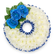 A bespoke funeral tribute shaped precisely as the number '0', shown from a soft top-down view against a clean white backdrop. The entire numerical form is built from hundreds of small, tightly clustered white spray chrysanthemums whose tiny, feathery petals knit together into a pillowy, tactile surface; the chrysanthemums give off a faint, comforting scent and a gentle matte sheen that reads as pure, consoling light. Around both the outer and inner rings of the zero there is a neatly pleated band of deep royal blue satin ribbon, folded into a scalloped trim that catches the light with a subtle satin glow and frames the shape with formal dignity. Nestled into the upper left curve is a focused cluster of three saturated royal blue roses, their petals unfurling from compact centers into velvety spirals; the blue is rich and slightly inky, a shade that suggests calm, respect, and a deliberate tribute to a favourite colour, regiment, or organisation. A few glossy dark green leaves tuck beneath the roses, adding a cool, botanical contrast and a living depth against the white chrysanthemum cushion. The overall effect is purposeful and serene - a personal, numbered memorial suitable for a commemorative service, wake, or graveside tribute. The composition balances softness and structure, the fluffy chrysanthemums tempering the formal ribbon border while the blue roses supply emotional punctuation. As a local florist we often prepare similar custom number tributes for families across Brent Cross and nearby Cricklewood, matching ribbon and rose tones to local choir or service colours, and offering careful same-day preparation to preserve freshness and respectful presentation.