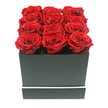 In this carefully composed photograph, a square arrangement of preserved red roses is presented with all the poise of a luxury gift, the kind you might send from Brent Cross Flowers for a memorable romantic occasion. At the centre of the frame sits a matte black box, perfectly square and smooth, its edges razor-sharp and its surface softly absorbing the light. Rising just over the rim of the box are twelve red roses arranged in an orderly 3x4 grid. Every bloom is at full openness, showing off tightly layered, velvety petals that curl inwards into rich, shadowed centres. The reds are deep and saturated, with slight tonal shifts that give life and movement to the otherwise uniform carpet of colour across the top. The container is kept deliberately simple, but is accented by a slim, bright white stripe that runs horizontally around its lower portion, adding a precise, contemporary detail. The scene is photographed against a completely white background, which removes all clutter and places the full focus on the interplay between the black box and the crimson roses. The lighting is soft and even, casting only gentle shadows that define the edges of the petals and the base of the arrangement, subtly emphasising depth without introducing drama. The overall effect is one of modern elegance and contained passion, making this design particularly suited to anniversaries, Valentine's Day, or a significant apology where words alone might not feel enough. It is easy to imagine this box of preserved roses gracing a hallway console or bedroom dresser in a Brent Cross home, serving as a daily reminder of a cherished connection.