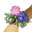 A modern-romance wrist corsage rests gently on a light-toned forearm, arranged to read like a small botanical jewel. At its center a velvety lavender-pink rose unfurls in a soft spiral, its petals edged with a slightly deeper magenta that catches the soft studio light and suggests the faint, cool scent of early summer blooms. Clustered close around the rose are rich indigo hydrangea florets, each tiny petal cradling a pale blue-to-ivory eye that gives the group a luminous, almost twilight-blue glow. Variegated ivy leaves frame the composition with classic dark green centres and creamy-yellow margins, their glossy surfaces reflecting a hint of sheen. Behind and between the flowers, feathery silvery-green sprigs-reminiscent of eucalyptus or lavender foliage-add a soft, muted backdrop, while a sculptural broad leaf with crisp white-and-green stripes is artfully curled into a tight spiral, introducing a contemporary, architectural note. Beneath the blooms a lacy, open-weave net in deep purple peeks through, its texture amplifying the hydrangeas' colour and lending a whisper of evening glamour. The whole piece reads as handcrafted craftsmanship: tactile contrasts from silky rose petals to the crisp ivy and the airy mesh invite a gentle touch. Intended for proms, bridesmaids, or a guest of honour, this wristlet carries the quiet joy of celebration and the freshness of a garden after rain. As a local Brent Cross florist I imagine it paired with a midnight-blue gown or a soft silver cuff, available for careful collection or local delivery across Brent Cross and neighbouring streets for those last-minute special moments.