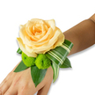 A delicate wrist corsage rests along the inner forearm, the composition seen in soft, evenly diffused light that highlights texture and tone. At its centre sits a single, fully opened peach-apricot rose, petals layered in a warm spiral that reads both tender and confident beneath the light. Tucked just to one side are two bright lime-green button chrysanthemums-small, spherical pom-poms whose densely packed florets create a playful, pebbled texture against the rose's satin-smooth petals. Broad deep-green leaves, likely salal or ruscus, form a lush cradle beneath the blooms, their glossy surfaces reflecting a hint of the studio light and giving the piece depth and weight. A folded, variegated leaf with crisp, lighter stripes sweeps in an architectural arc from beneath the rose toward the wrist's edge, lending a modern, ribbon-like line that balances the rounded flowers. The corsage is styled to sit flush with the skin, the fastening discreet and hidden so the flowers appear to float; the skin tone visible is a warm, light to medium brown that gently warms the palette. The scent, imagined from fresh roses and cut greenery, would be a faint, clean rose note with a green, herbaceous undercurrent-perfect for intimate vows or a prom evening. As a local florist I picture this worn on a walk between Brent Cross Shopping Centre and Cricklewood Broadway, or while arriving for photographs in nearby Gladstone Park, offering refined, wearable beauty for community celebrations and neighbourhood ceremonies near Brent Cross.