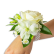 A delicate white wrist corsage photographed against a pure white backdrop, the wearer's light skin and wrist angled from the bottom right toward the centre to showcase the arrangement. At its heart sits a creamy ivory rose, petals unfurling in a soft, velvety spiral that catches the light with a gentle, almost pearlescent sheen. Below and slightly left of the rose a larger, trumpet-shaped white bloom - suggestive of an Alstroemeria or Peruvian lily - opens wide to reveal a pale green-yellow throat that adds subtle depth and a natural focal contrast. Interspersed around these central flowers are fresh green buds, some tightly closed and promising new life, while airy sprays of Baby's Breath (Gypsophila) lend a cloud-like, whispery texture. Near the wrist a cluster of small daisy-style white florets with bright yellow centres brings a cheerful, delicate punctuation to the design. Broad, glossy deep green leaves frame the composition, their smooth, waxy surfaces giving the whites a lush backdrop. The whole piece is secured with an elegant multi-strand pearl bracelet; dozens of round, lustrous white pearls wrap gently around the arm, the cool smoothness of the beads contrasting softly with the petals' tender give. Under soft, diffuse studio light the colours read as clean and refined, and there's a faint, fresh floral scent - powdery rose with a hint of green. Perfect for prom, wedding, or formal occasions across Brent Cross and nearby Hendon, this wristlet balances classic sophistication with a handcrafted, garden-fresh feel.